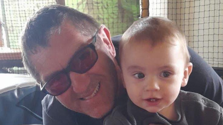 A close-up photo of faces, showing Leon, aged 56, holding his grandson, Michael, who appears to be sitting on his lap. Leon is leaning forward putting his head beside Michael's, and smiling at the camera. He has grey hair and is wearing sun glasses. Baby Michael has wispy light-brown hair and two bottom teeth. 