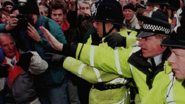 Three police officers wearing hi-vis jackets trying to control crowds of protesters.