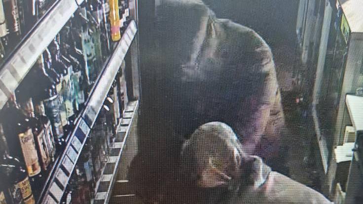 CCTV footage of two people in hoods and coats next to an alcohol shelf