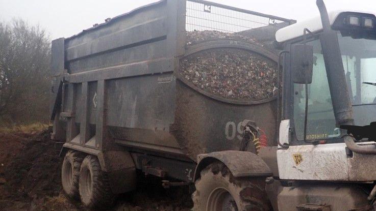 A white JCB tractor with a trailer that contains waste. 