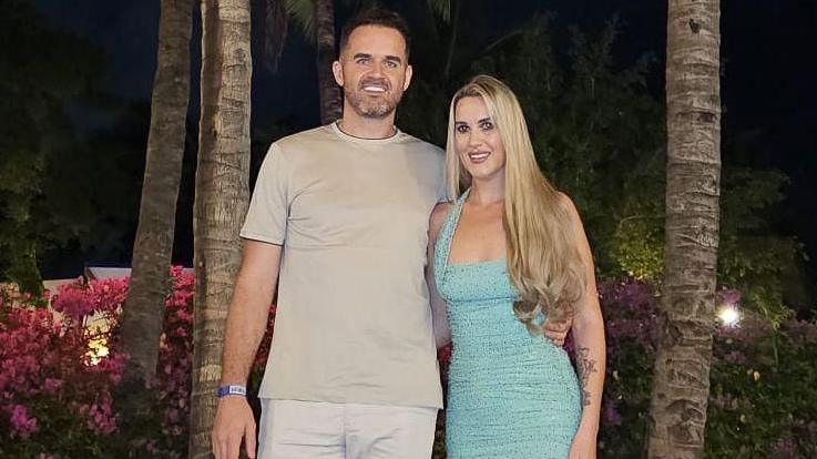 Liam is wearing a beige t-shirt and beige shorts and Natasha is wearing a light blue dress. They are standing in front of palm trees at night time.