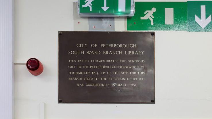 A brown plaque put up on a wall says: "CITY OF PETERBOROUGH SOUTH WARD BRANCH LIBRARY. This tablet commemorates the generous gift to the Peterborough corporation by HB Hartley ESQ JP of the site for this branch library, the erection of which was completed in January 1951".