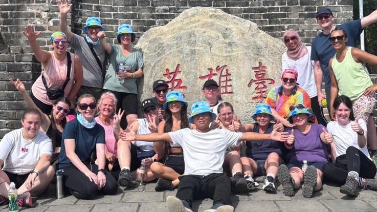 A group of people stand by a large stone with chinese writing. They wear walking gear and hats and sunglasses.
