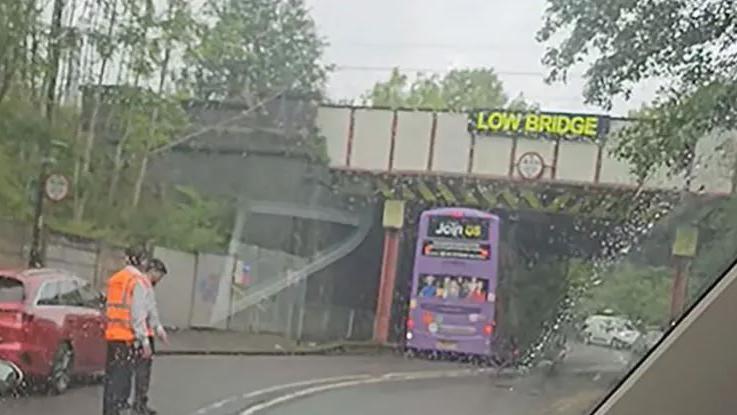 A purple double decker bus has crashed into a low bridge with two people in orange hi-vis sleeveless vests standing behind it and parked cars behind it. It is raining.