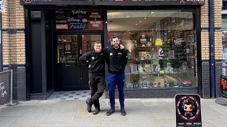 Image of a man with his arm around the shoulders of another man in front of their shop. There are collectable toys in the shop window.