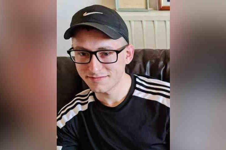 Martyn wears a black sports T-shirt, a black sports baseball cap and black-framed glasses. He is cleanly shaven and is sitting on a brown sofa inside.