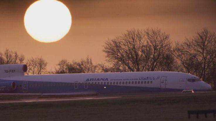 A picture of an Ariana Airlines flight at Stansted Airport with the rising sun in the background.