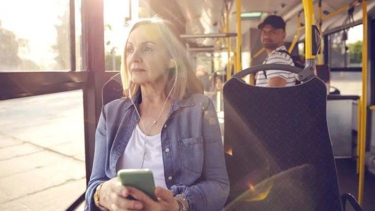 A woman wearing a purple cardigan is listening to her phone through earplugs on a bus. A man in a cap is looking in her direction.