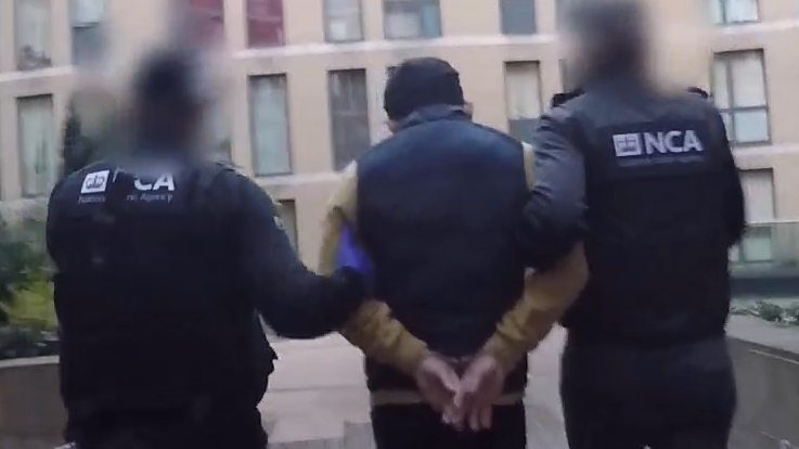 Le is led away from his flat by two NCA officers in uniform. He has his hands cuffed behind his back and his back to the camera as do the officers.