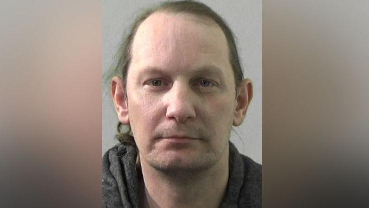 Mugshot of Winters. He is clean shaven with long straggly brown hair and wearing a dark grey hooded top.