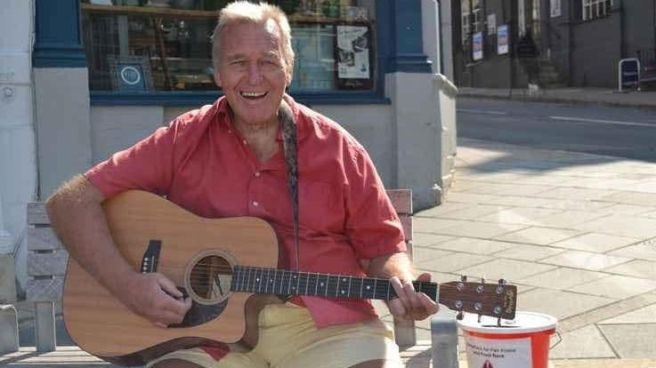 Paul busking with his guitar to raise money for local charities, sitting beside a collection bucket 