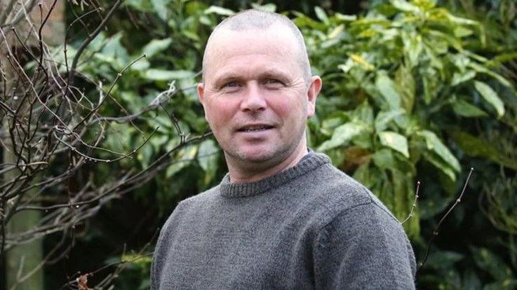 Dessie Loughery, who is bald, wearing a grey woollen sweater and looking directly at the camera. He is stood in front of a hedge