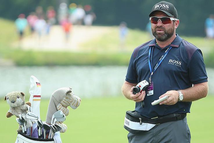 Craig Connelly on a golf course. He is wearing a blue Boss T-shirt, Boss baseball cap and sunglasses. He is carrying a camera and has a bum bag around his waist. He is standing next to a set of golf clubs, some of which have animal covers on them