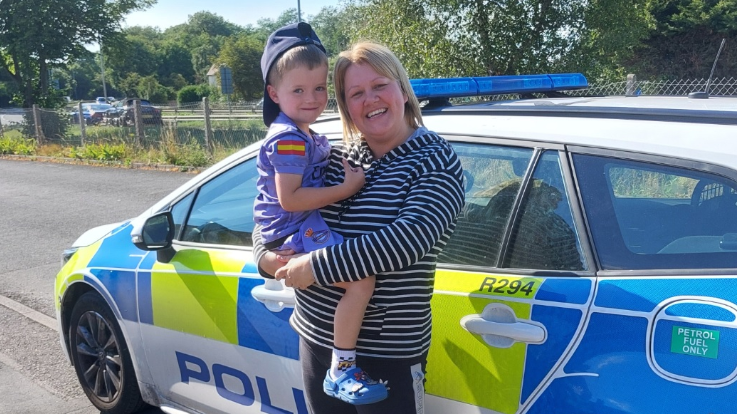 Pictured is a mother and young boy, her son in his mother's arms, stood in front of a British white, blue and yellow police car smiling. The little boy has blonde hair and is wearing a purple hat backwards, a purple t-shirt and blue crocs. His mum has short blonde hair and is wearing a black and white stripe jumper with a pair of black Adidas leggings with a grey stripe along the line.