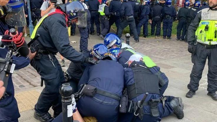 A man lying on the floor with five police officers on him, wearing riot gear and helmets, while a crowd surrounds them.