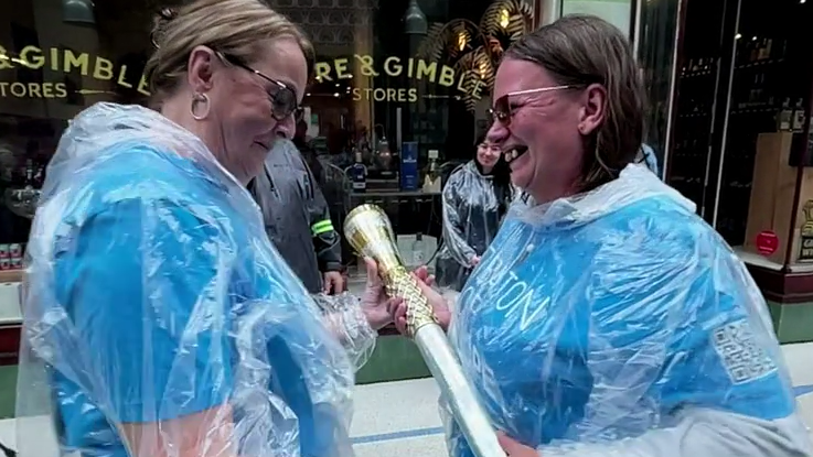 Two women standing side on to the camera and facing each other. The one on the right, who has brown hair and is earing glasses, is handing a silver and gold baton to the one on the left, who has lighter brown hair and sunglasses. Both are smiling and wearing blue tshirts with clear plastic rain covers over the top.