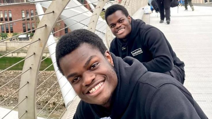 James and John Makanjuola are outside on a bridge, cleaning. They are both looking at the camera and smiling, with one behind the other and wearing matching black hoodies and tracksuit trousers.