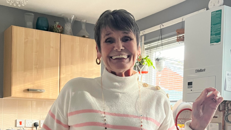 A woman with short grey hair wearing a pink and white jumper. She is standing in front of cabinets in a kitchen and is smiling to the camera. 