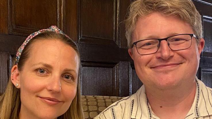 A young woman on the left with a flowery headband. Paul Mitchell is on her right, he has grey hair, a white striped shirt and square glasses. They are both smiling into the camera. They appear to be in a pub.