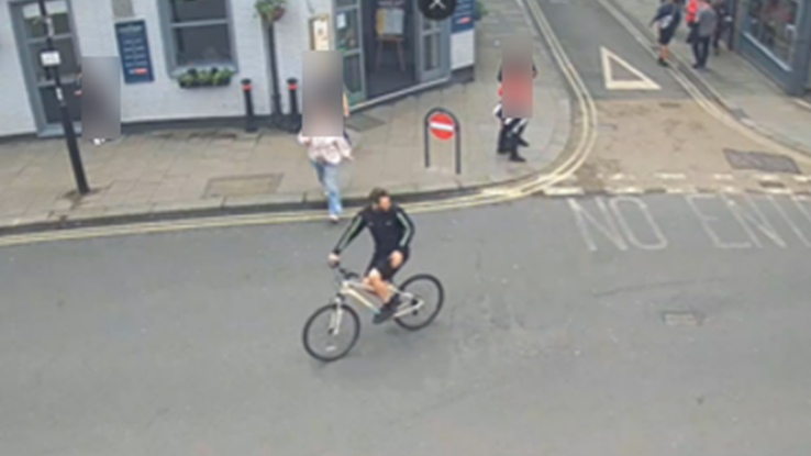 A street scene captured by CCTV shows a cyclist riding towards the camera. On the corner of the street is a pub and pedestrians are blurred from view.