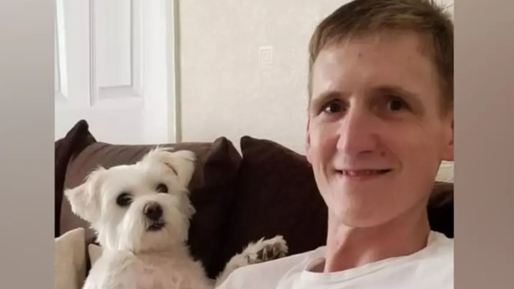 A man with light brown hair and wearing a white t-shirt sits on a brown sofa with a white terrier dog sitting next to him.