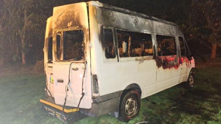 A white minibus seen from the back and side. The windows are all gone and the top half looks burned out. It is parked on grass by trees. By the back doors there is a wheelchair ramp, folded up.