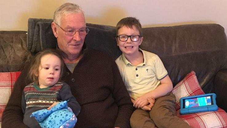 The photograph shows an elderly man wearing glasses and a brown jumper, sitting on a sofa with two children. A little girl is sitting on his lap and a boy is sitting by him smiling at the camera. 