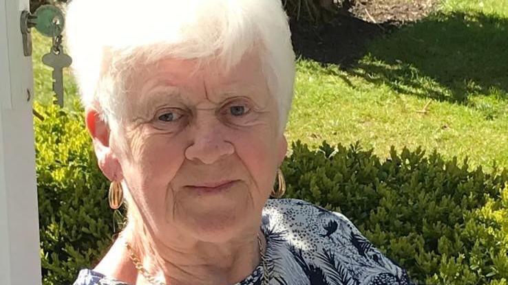 Marie Cunningham with white hair and gold hoop earrings wearing a black-and-white patterned blouse. She is sitting in a garden.