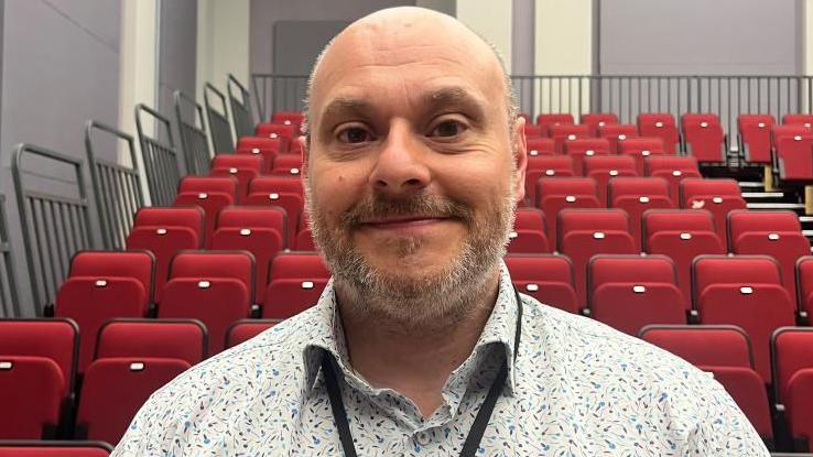 Lee Johnson is looking into the camera, smiling. He has a shirt, grey beard. He is wearing a white and blue patterned shirt and a black lanyard round his neck. Behind him, there are red pull down theatre seats.
