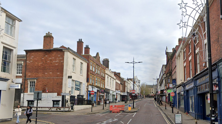 The street has shops on either side. A barrier on the left hand side of the street prevents traffic from driving on to it.