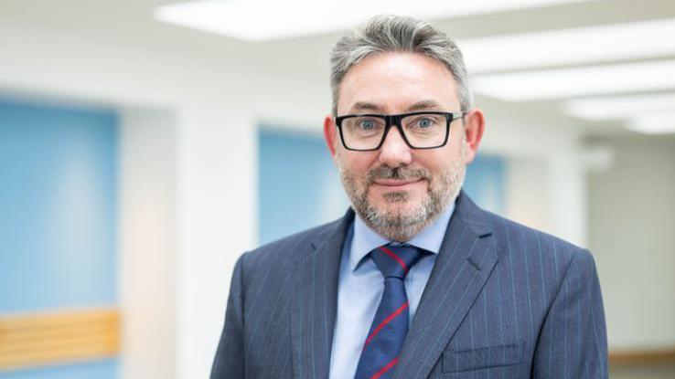 A man with grey hair and a grey beard. He's wearing glasses and a blue stripe suit with a blue shirt and a blue tie with red stripes.