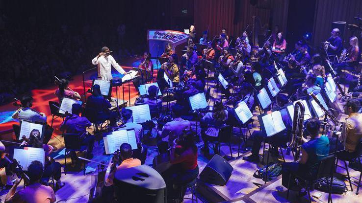 A conductor dressed in white leads a large orchestra. The picture is taken from behind the musicians, so you can see their open sheet music, and they are lit by dark blue and purple lighting. 