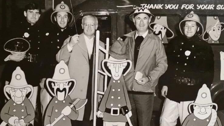 A black and white photograph of five men, standing in front of four cut-outs of cartoon firefighters. Three are in firefighter gear, wearing dark, buttoned jackets with a large belt across the torso, light-coloured trousers, and a light-coloured helmet. The two other men are dressed in civilian clothes.