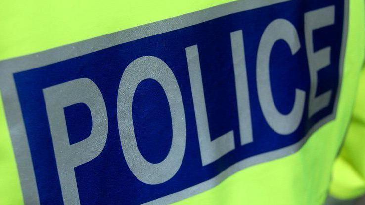 Close up of the back of a police officer's high visibility jacket. The word "police" is in silver letters on a blue background.