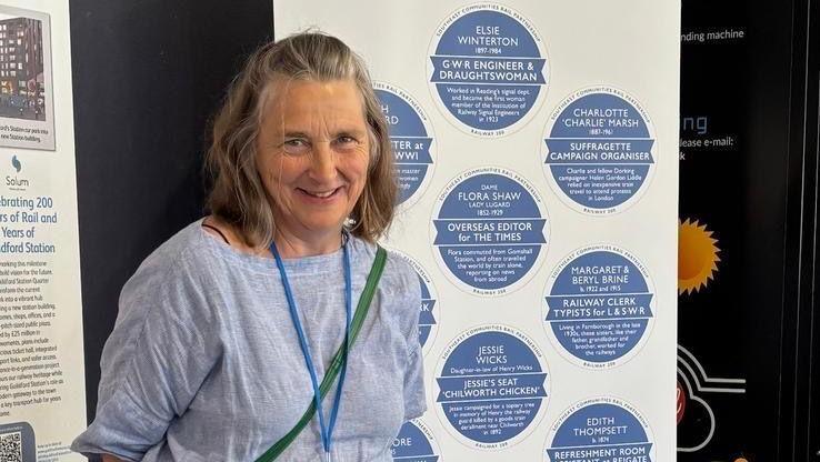 Sara Grisewood stands in front of a banner showcasing the names of 10 women who contributed to the history of women's involvement to the railway line. She is smiling at the camera wearing a blue shirt and lanyard. Visible names on the banner include Elsie Winterton, Charlotte 'Charlie' Marsh, Dame Flora Shaw, Margaret and Beryl Brine, Jessie Wicks and Edith Thompsett.