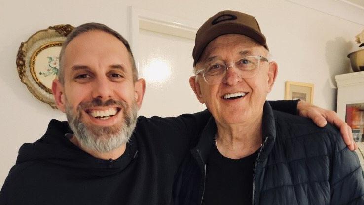 A smiling, bearded Matthew with his arm around his father, who is also smiling, wearing glasses and a cap. Both are dressed in black.