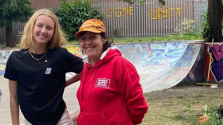 A young woman with shoulder-length blond hair, wearing a black t-shirt branded with "SB GB" stands smiling next to an older woman wearing a brightly coloured baseball cap and a red hoodie. Both are smiling at something behind the camera and are standing in front of a skateboard ramp which has been decorated with graffiti.