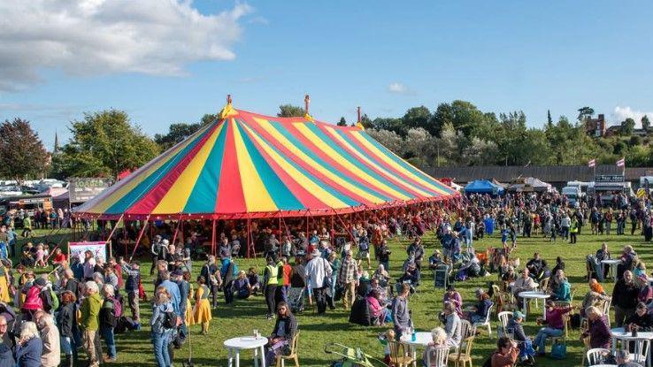 A large multi-coloured tent, in a field with lots of people stood beneath and around. The ten seems to be in a park, with trees and buildings in the distance.