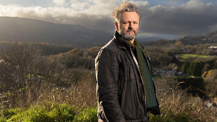 Standing on high ground above a valley, Michael Sheen looks towards the camera with rolling hills and patchwork fields stretching into the distance. He is wearing a brown jacket with a green patterned jumper. Sheen has grey hair and facial hair. Strong afternoon sunlight and heavy clouds create dramatic contrasts across the rural landscape.