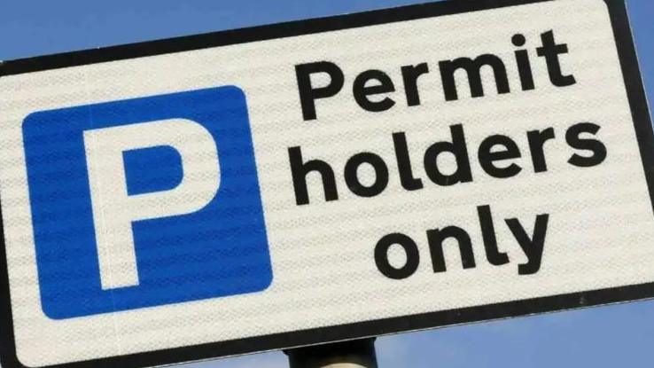 A white sign that says Permit holders only, the writing of the sign is black, it has a blue box with a white P in it, and there is a large blue sky behind the sign. 