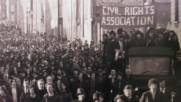 A black and white photograph of hundreds of people marching along a street in Londonderry on 30 January 1972.
On the right hand side of the image is a truck, with people standing on the back holding a large sign which says Civil Rights Assocation.
