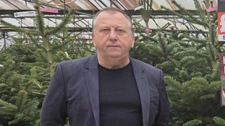 Plants Galore owner Tony Joyner stands in front of dozens of Christmas trees at a shop in Newton Abbot. He is wearing a dark blue blazer and black t-shirt. He has a stern look on his face.