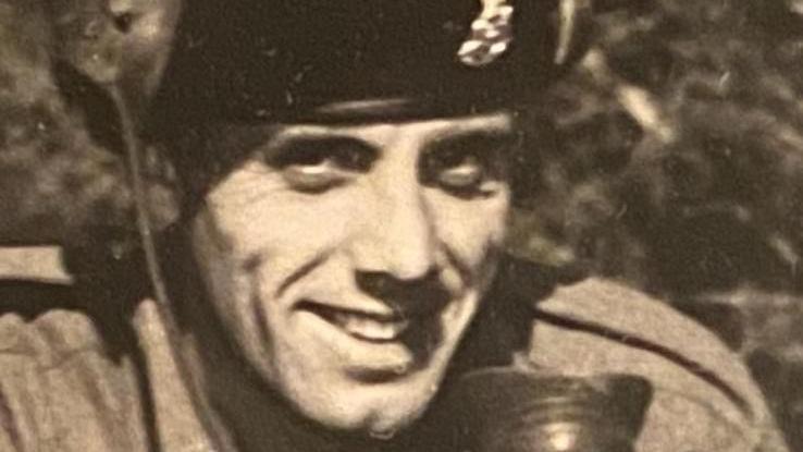 A black and white picture of a man in military uniform. He is smiling at the camera and has a badge on his beret