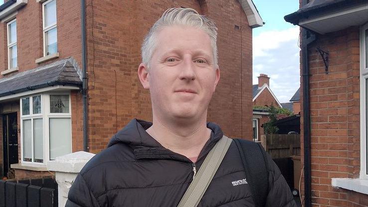 Craig McConnell smiling at the camera. He has short white/grey hair and is wearing a black hooded coat. He is standing in front of red-brick houses. 