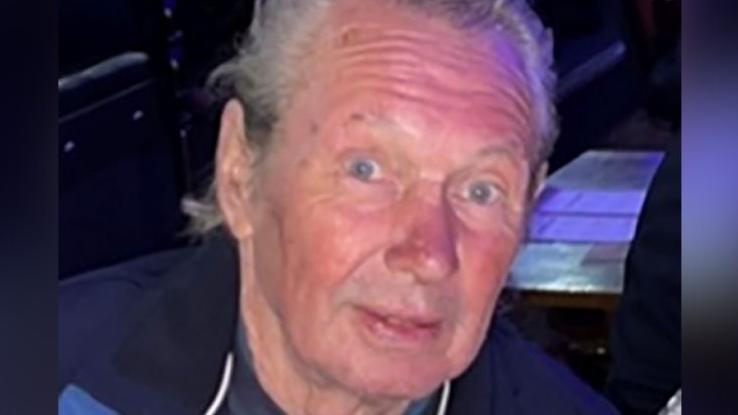 Peter Eric Greener is smiling. He has short grey hair and is wearing a grey top and blue coat.