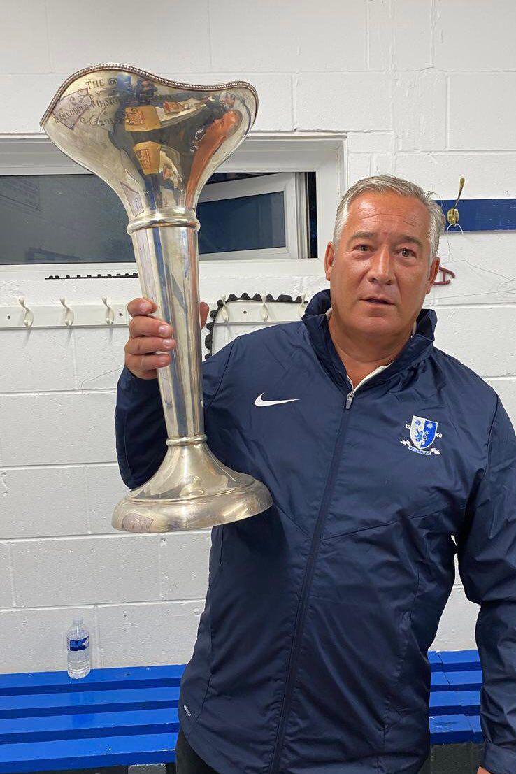 Richard Pillinger, who has grey hair and is wearing a blue tracksuit jacket. He is holding a silver trophy.