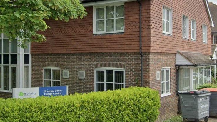 A tile and red brick building with white window frames behind a green hedge. A sign reads "Modality: Crawley Down Health Centre"