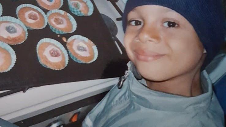 Ty’rellé Burke, wearing a blue woollen hat and a grey zip-up raincoat, smiles at the camera next to a tray of cupcakes 