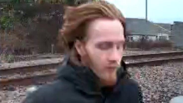 A man with ginger hair and beard, wearing a dark jacket, is running across a railway track. His face is slightly blurred and pixelated. Metal railings run alongside the track, and the background features railway lines, vegetation and distant buildings.