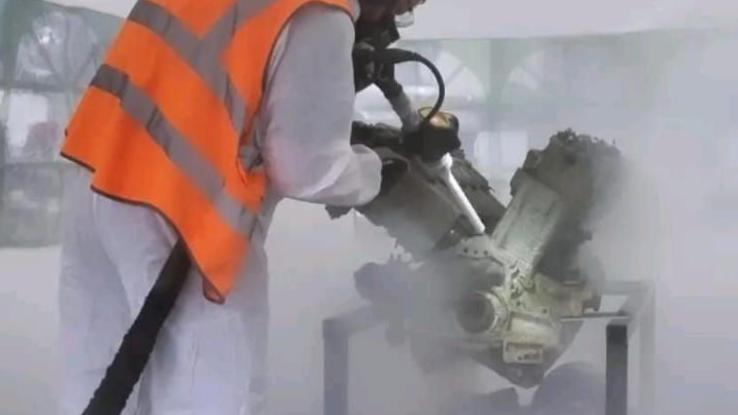 A man wearing a full white hazmat suit and orange high vis vest and a mask, leans over an engine and blasts it with a large piece of dry ice equipment. Dry ice is pictured swirling around. The cleaning is happening inside a large white tent structure.